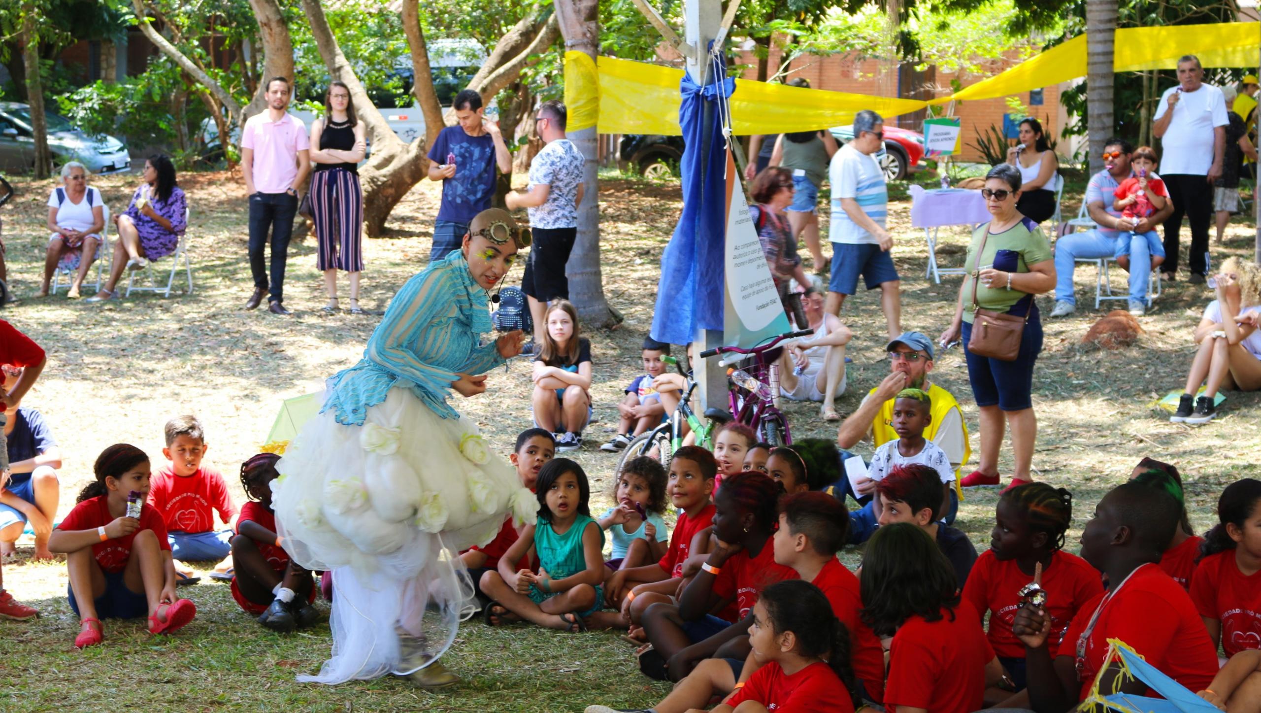 Crianças em roda assistem a uma pessoa fantasiada se apresentar numa praça arborizada, enquanto adultos também observam ao fundo.