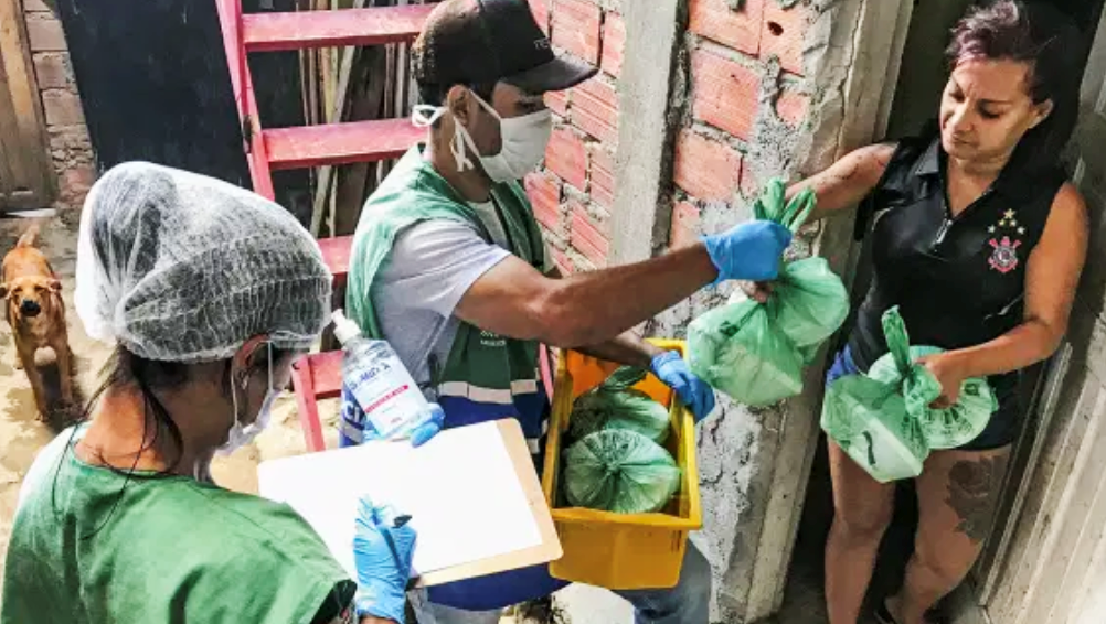 Foto mostra dois voluntários entregando sacolas com mantimentos para uma moradora em Paraisópolis, em São Paulo