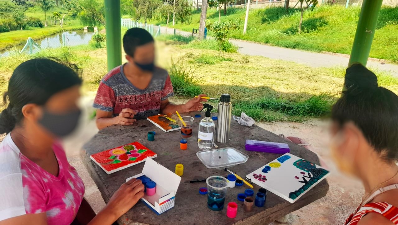 Duas mulheres e um homem estão sentados ao redor de um mesa, em um ambiente externo. Sobre a mesa estão tintas, pincéis e telas de pintura.
