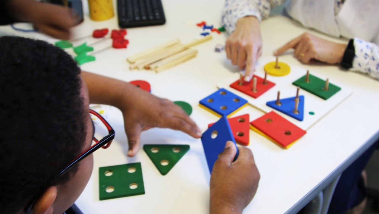 Imagem mostra uma criança participando de uma atividade pedagógica, que inclui diversas peças em formatos geométricos espalhadas na mesa.