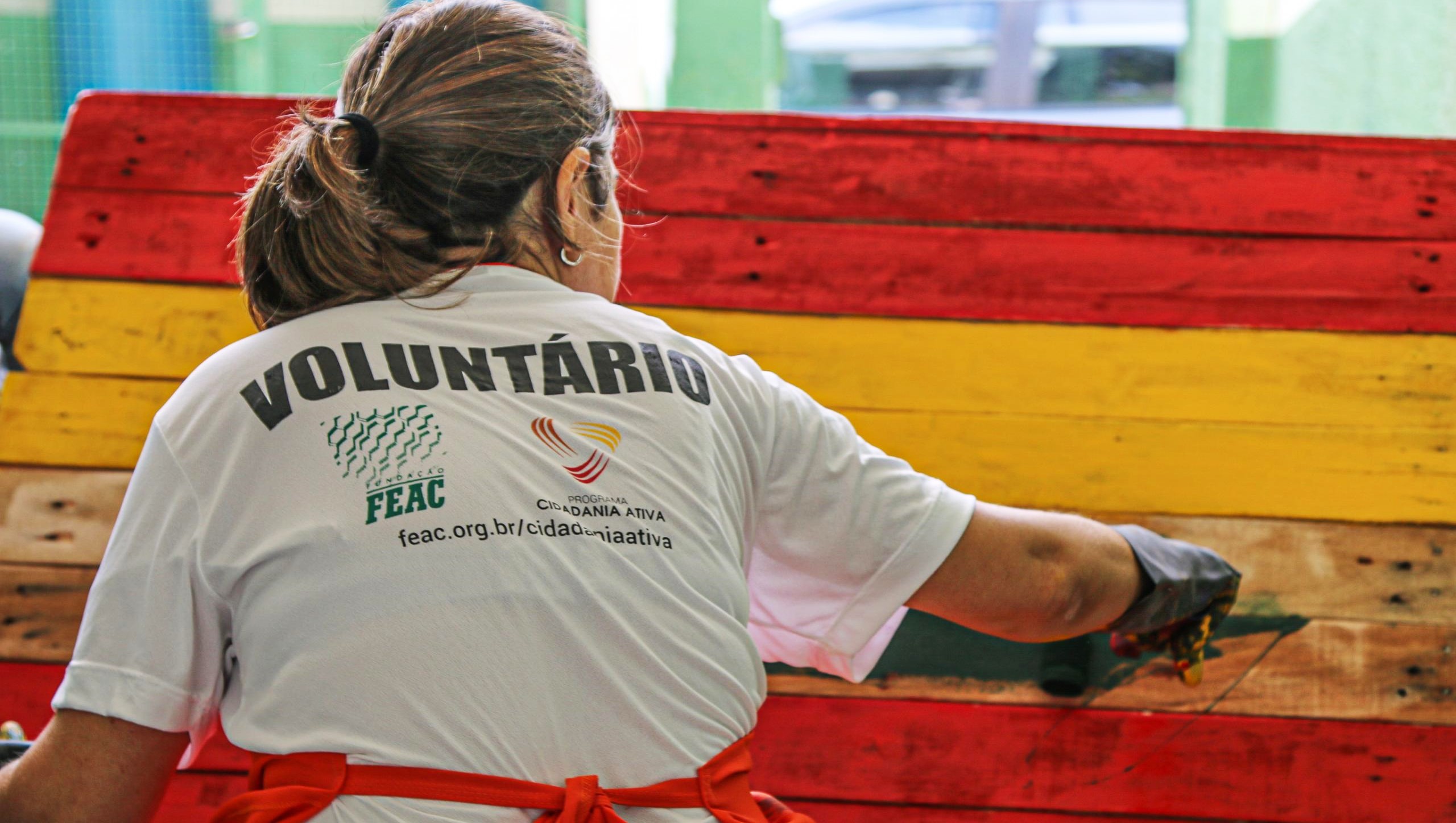 Foto exibe mulher de costas pintando de vermelho e amarelo um objeto de madeira. Na sua camiseta está escrito "voluntário".