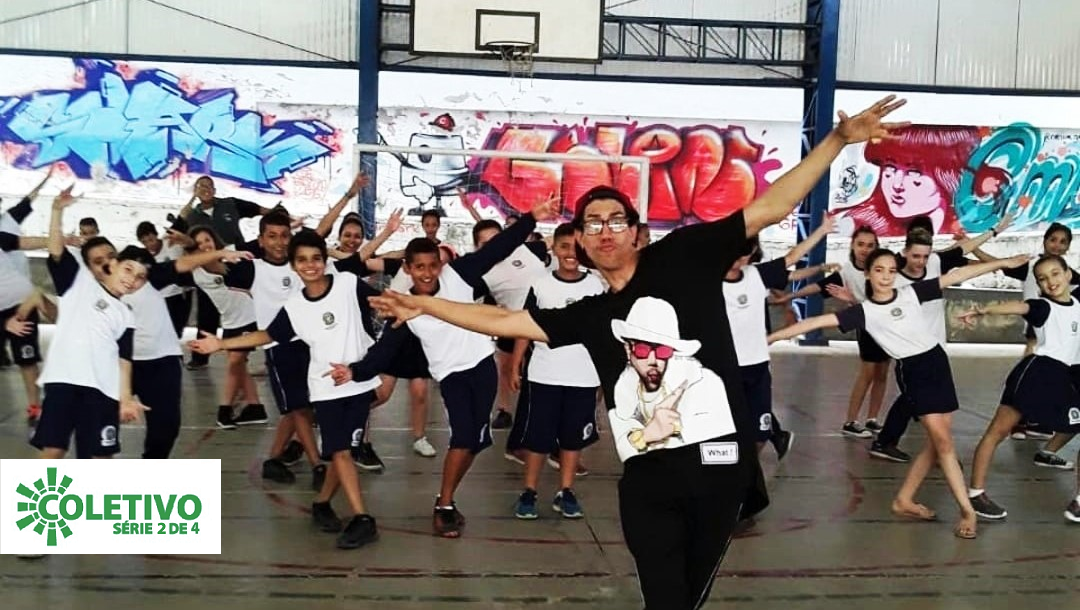 Imagem mostra um integrante do coletivo P2RCA à frente de várias crianças com uniforme escolar. Eles estão posando para a foto, com os braços abertos, na quadra da escola.