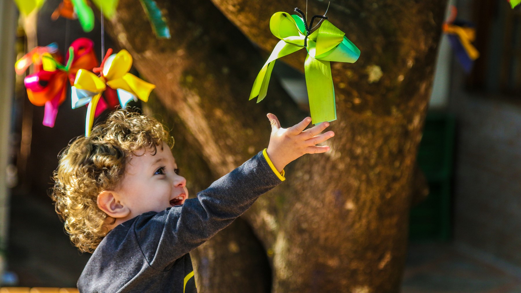 criança sorridente tenta alcançar um laço verde pendurado em uma árvore.