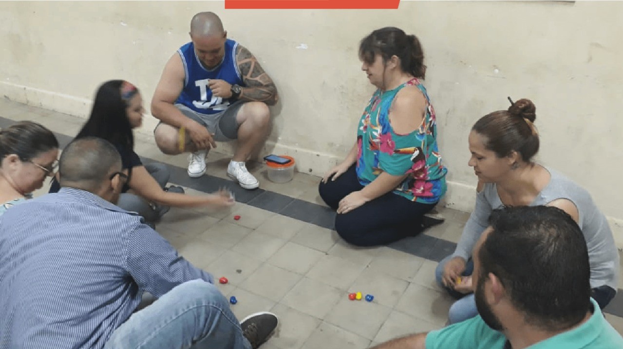 Sete pessoas estão sentadas no chão, fazendo uma atividade com pequenas bolas coloridas durante uma capacitação para famílias que queiram ser acolhedoras.