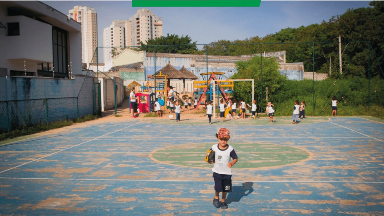 Em uma quadra ao ar livre, uma criança pequena, usando boné e uniforme branco, vai em direção à câmera. Ao fundo, há mais crianças brincando.