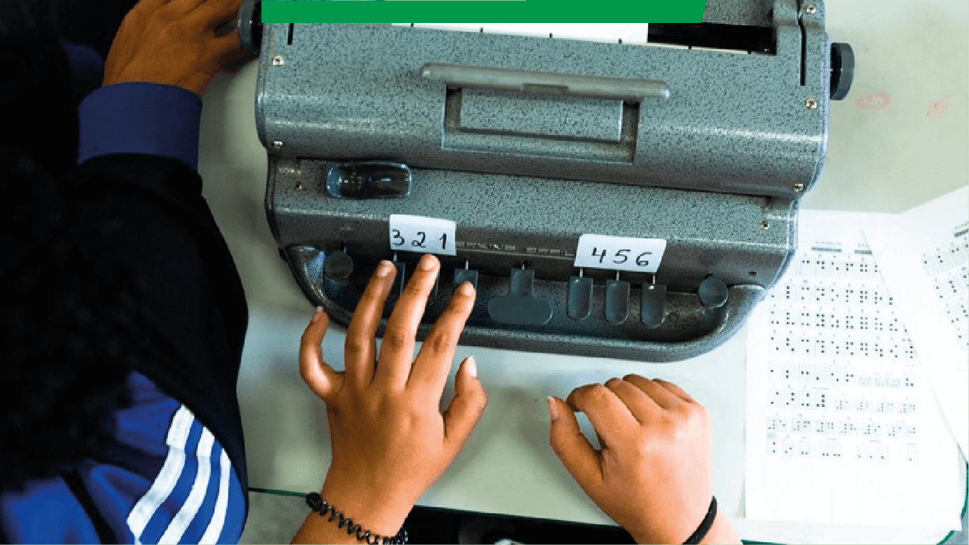 Imagem mostra, de cima, uma máquina de escrever especial, que escreve em Braille. Duas mãos estão na máquina. Do lado direito, duas páginas digitadas em Braille.