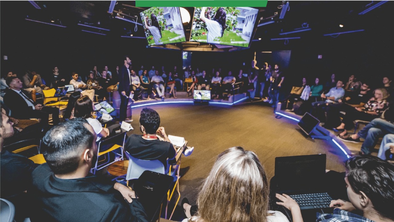 Foto exibe um pessoas sentadas em uma sala de eventos, com grandes monitores suspensos no centro do ambiente, durante encontro da Fábrica de Startups