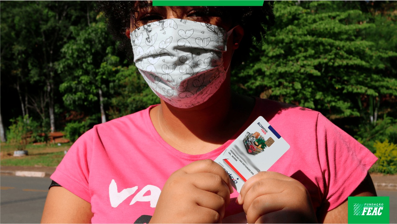 Foto de uma mulher segurando um cartão alimentação do Mobiliza Campinas, de frente para a câmera. Ela usa máscara e camiseta rosa.