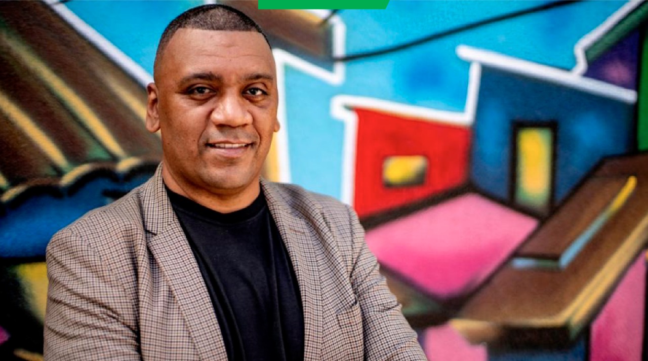 Foto de Preto Zezé. Ele é um homem negro, de cabelo raspado, está sorrindo de braços cruzados, usando um blazer quadriculado, posando na frente de um muro colorido grafitado.