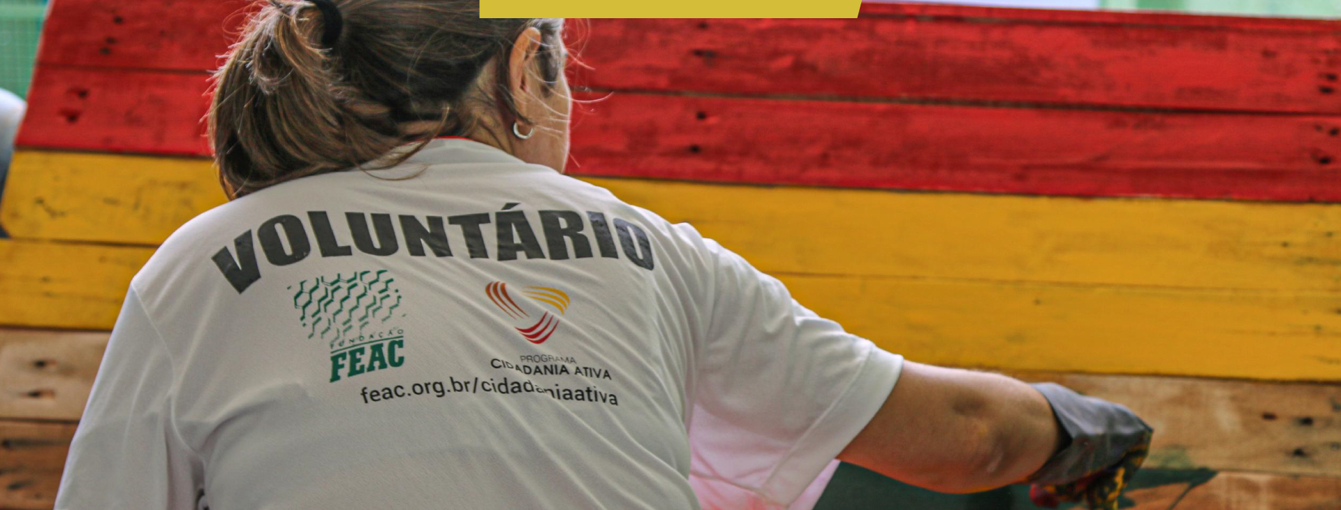 Mulher virada de costas usando camiseta escrita "voluntário" pinta painel de madeira nas cores vermelho, amarelo e verde