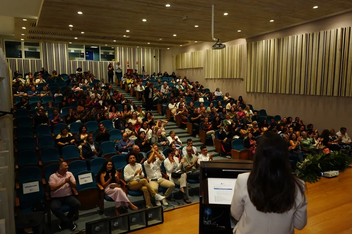 Vista panorâmica de auditório lotado durante cerimônia de certificação da Academia Social, com plateia aplaudindo. Ao fundo, palestrante de costas em pé na tribuna com notebook. Cadeiras azuis e decoração com plantas ao lado do palco.
