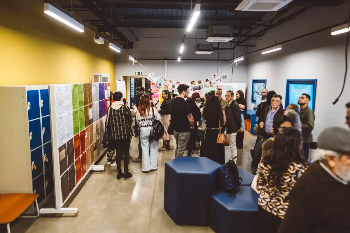 Participantes circulam por exposição de trabalhos durante cerimônia de certificação da Academia Social, observando painéis coloridos fixados em suportes ao longo de corredor. Ambiente moderno com paredes amarela e branca.