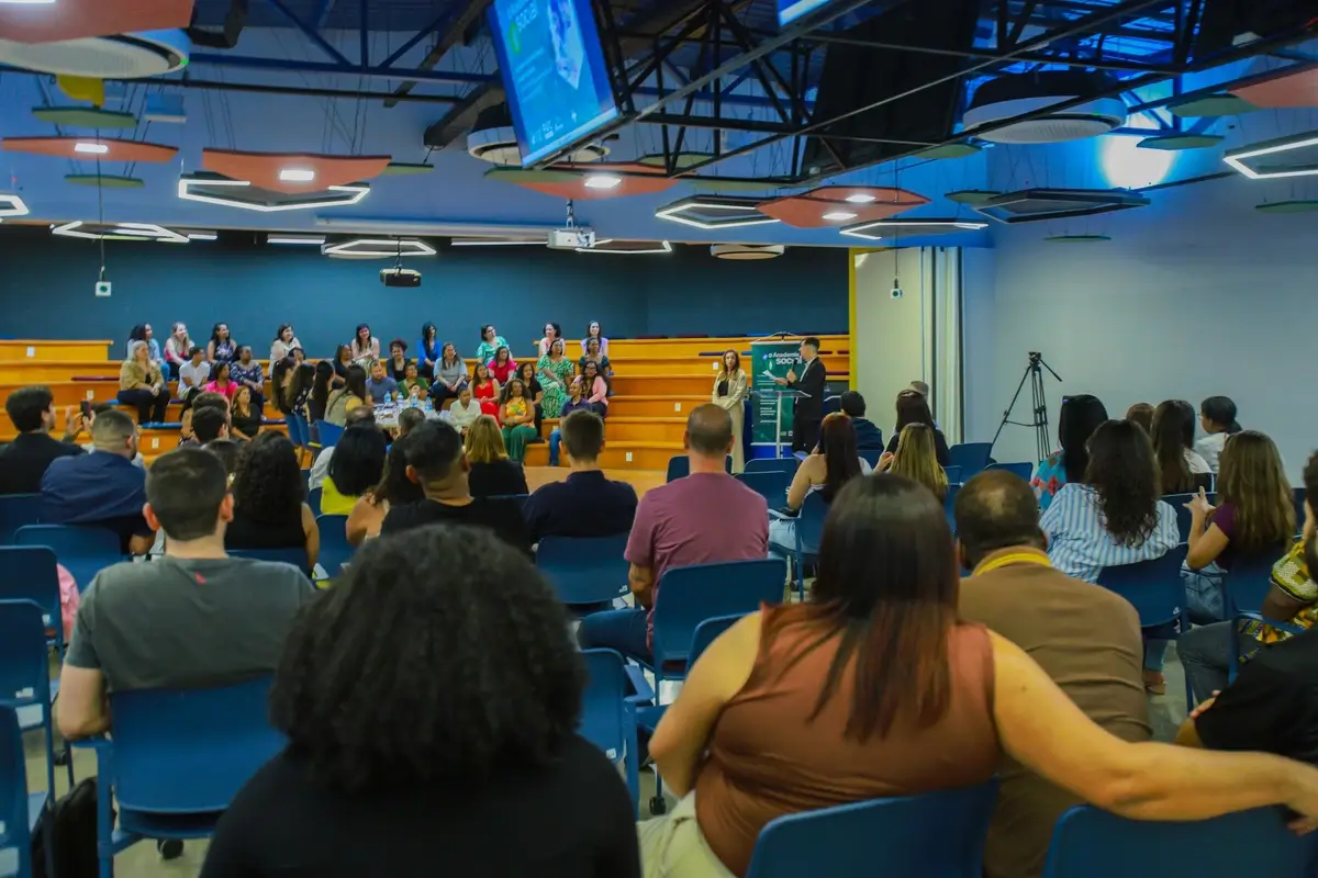 Turma certificada posada nas arquibancadas do palco durante cerimônia da Academia Social, com plateia sentada à frente e banner verde da Academia Social ao lado da tribuna. Auditório moderno com iluminação colorida no teto.