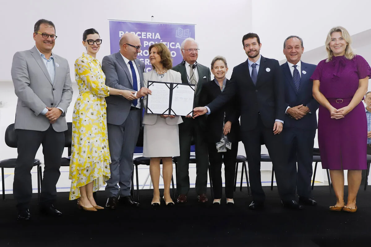 Grupo de nove pessoas posadas em palco durante cerimônia de assinatura do Protocolo de Intenções entre Fundação FEAC, Prefeitura Municipal de Campinas e Childhood Brasil, com presença da Rainha da Suécia. Ao fundo, banner com texto "Parceria pela Política de Proteção Integral à Criança".