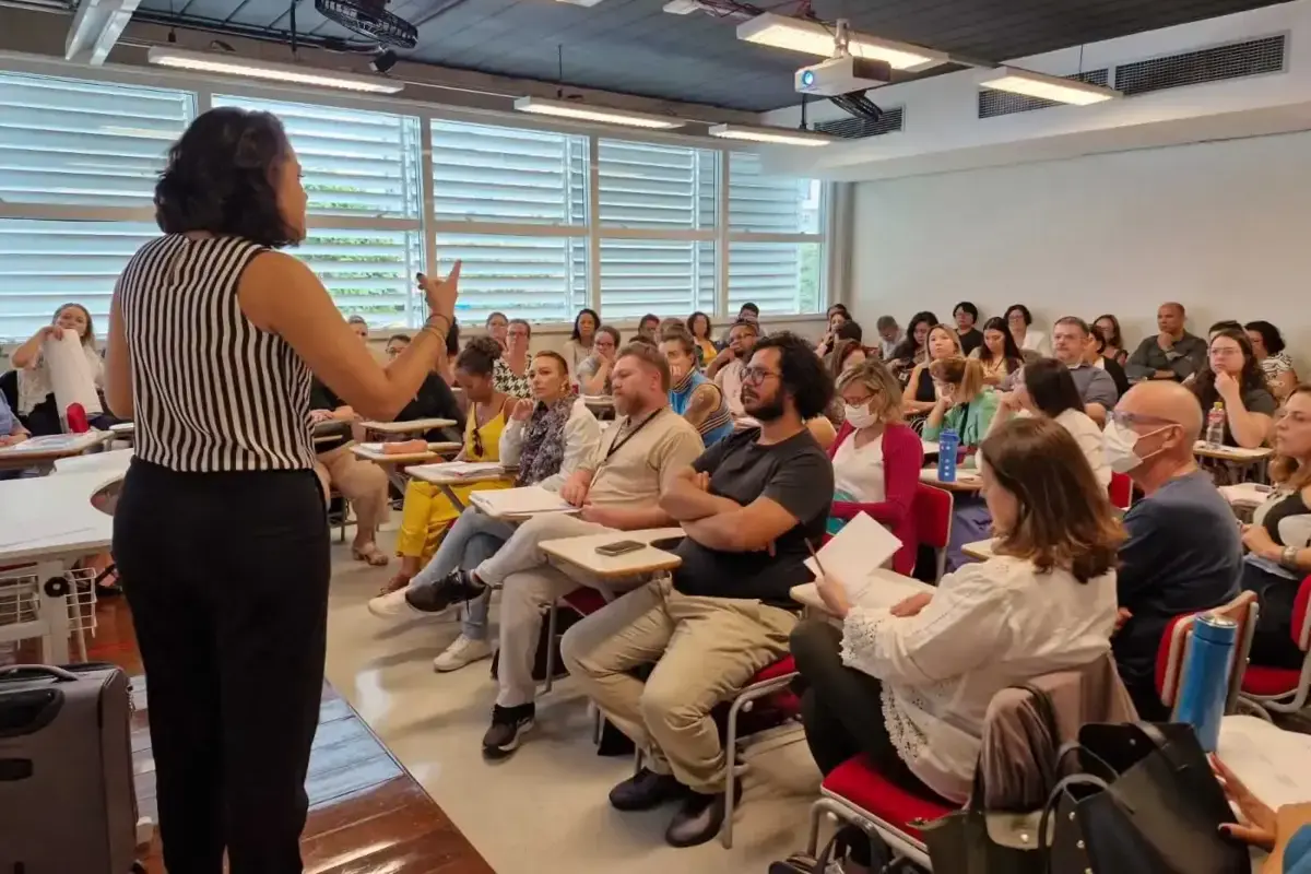 Palestrante de costas, com blusa listrada preta e branca, apresenta conteúdo gesticulando para plateia de cerca de 50 profissionais sentados em carteiras universitárias, durante oficina do CAI sobre a Lei Escuta Protegida.