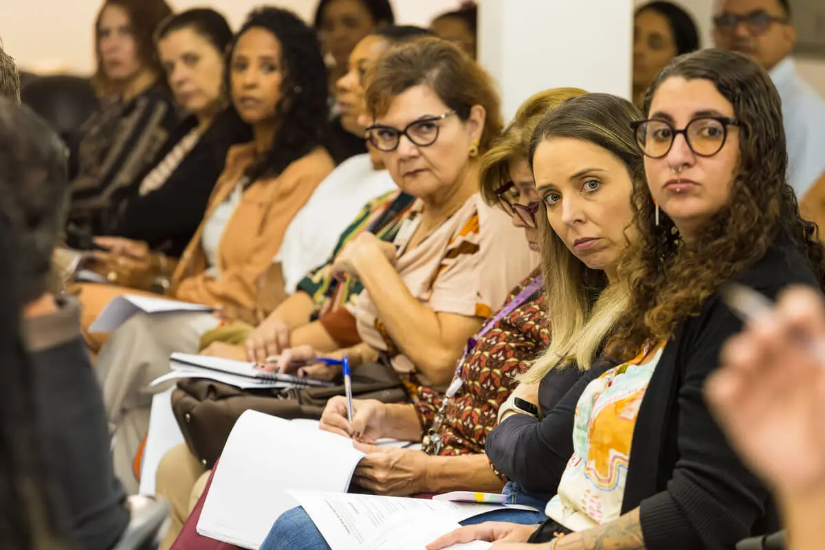 Grupo de participantes, majoritariamente mulheres, sentados em fileira durante oficina do CAI, com cadernos e documentos nas mãos, demonstrando atenção e engajamento na formação sobre a Lei Escuta Protegida.