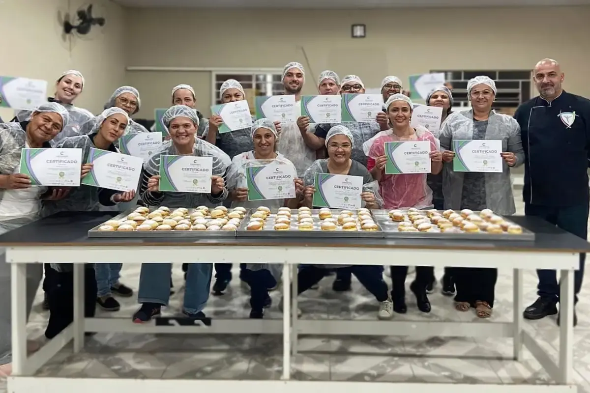 Grupo de pessoas em uma oficina culinária segurando seus certificados de conclusão e bandejas com salgados sobre uma mesa.