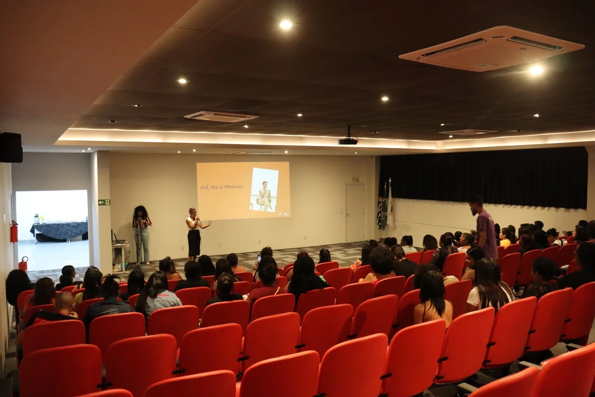 Foto interna de um auditório com poltronas vermelhas ocupadas por um público atento. No palco, uma mulher segura um microfone e fala para os presentes; ao fundo, um telão projeta a frase 'Oiê, sou a Mariana' acompanhada de uma imagem.