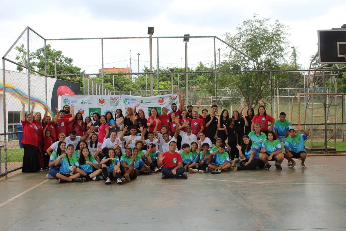 Foto horizontal de um grupo grande de pessoas reunidas em uma quadra poliesportiva durante o Festival de Ideias do projeto Hub de Cidadania Ativa. Muitos participantes vestem camisetas coloridas e fazem gestos com as mãos para a câmera, celebrando a realização do evento.