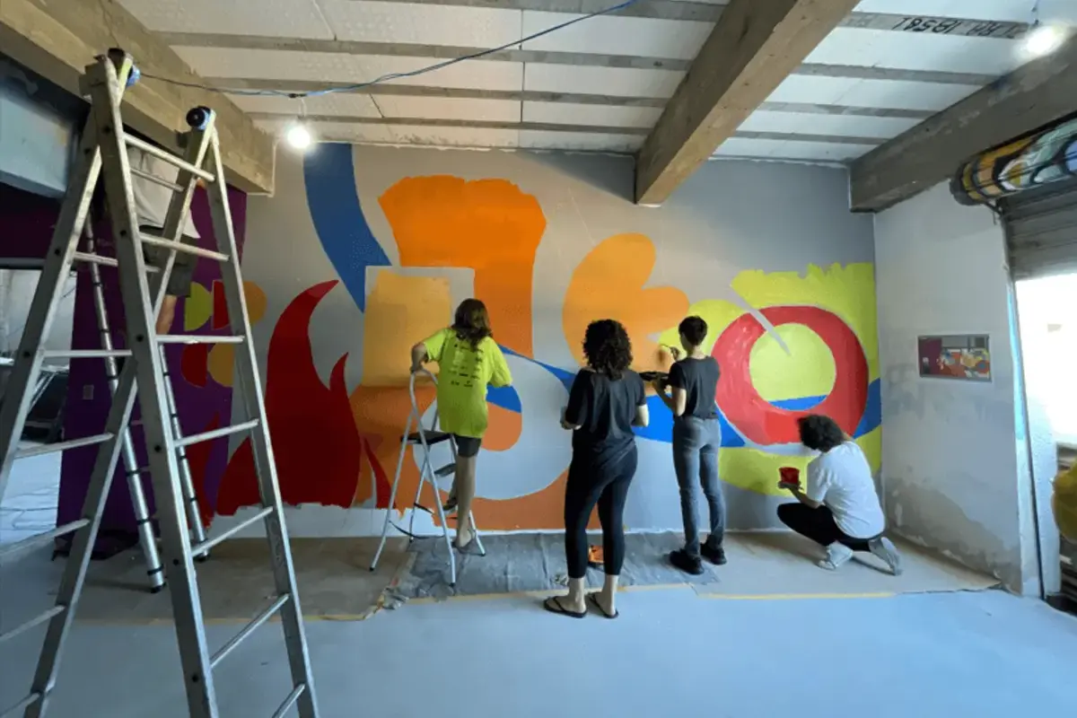 Foto horizontal mostrando um grupo de quatro pessoas trabalhando juntas na reforma e pintura de uma parede interna. Elas utilizam escadas e rolos de tinta para criar um mural colorido com formas abstratas em tons de amarelo, vermelho e azul.