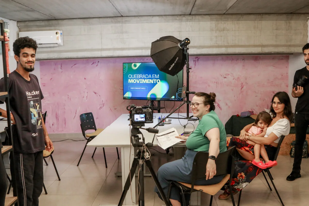 Foto horizontal mostrando pessoas do projeto Hub de Cidadania Ativa. Em uma sala, um grupo de pessoas participa de uma gravação de vídeo; há uma mulher rindo ao centro, uma mãe com sua filha no colo, um jovem em pé à esquerda e equipamento de filmagem, incluindo tripé e iluminação, no centro da sala. Ao fundo, uma TV exibe o logo 'Quebrada em Movimento'.