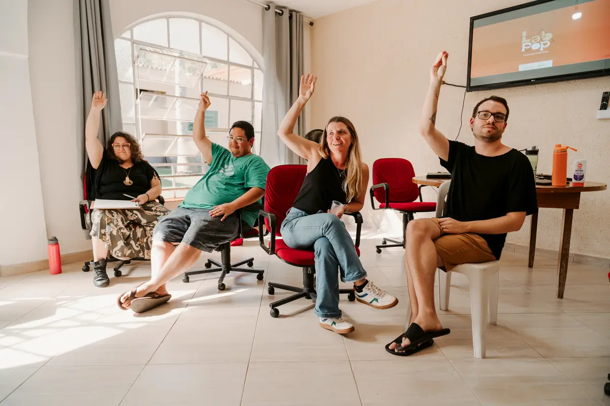 Quatro participantes do projeto LabPOP sentados em círculo com as mãos levantadas, participando de uma votação ou dinâmica.